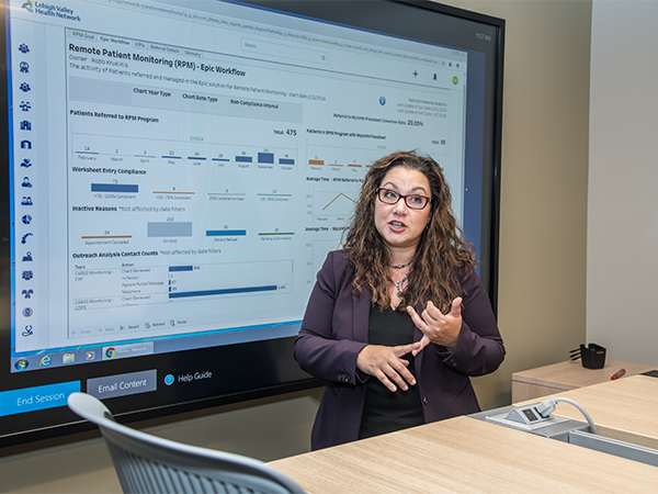 An adult stands in front of a digital presentation of data and information.